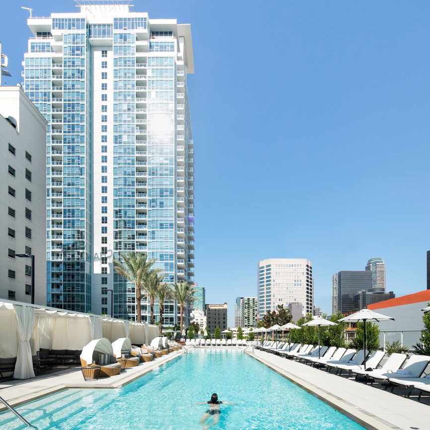 A person swims alone in a long outdoor pool lined with lounge chairs and cabanas, surrounded by tall modern skyscrapers under a clear blue sky.