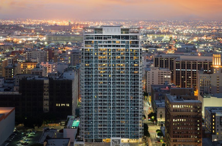A tall, modern high-rise building stands in the center of an illuminated cityscape at dusk, with city lights stretching into the distance and a colorful sunset sky above.