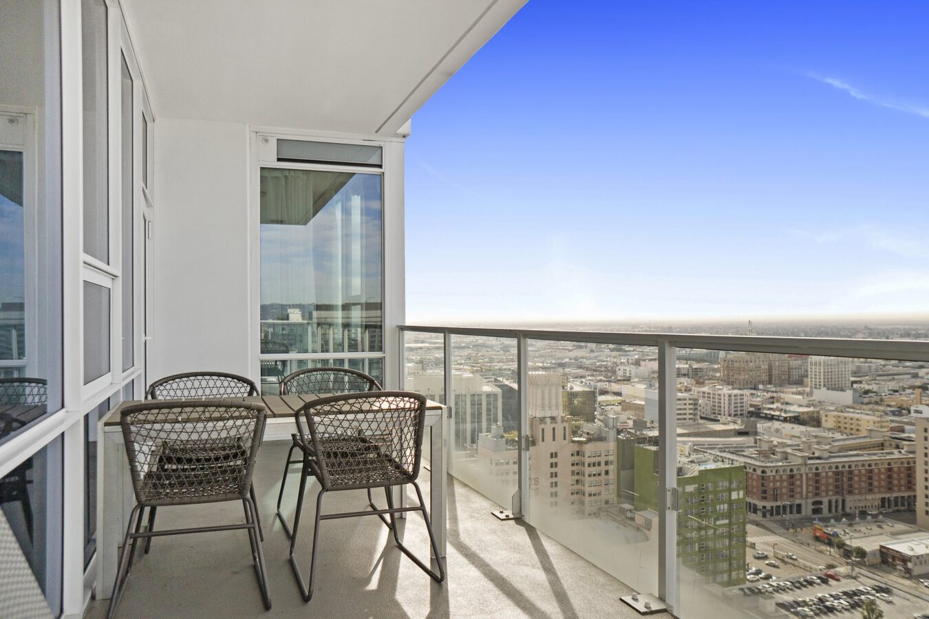 A modern high-rise balcony with a glass railing, a table, and four metal chairs overlooking a cityscape under a clear blue sky.