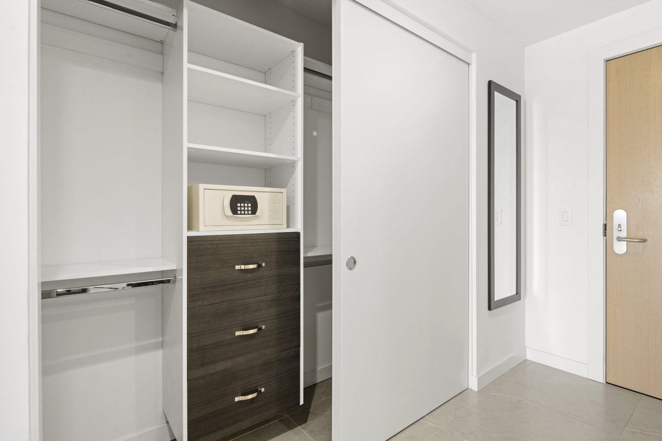 A modern closet with open shelves, a built-in safe, dark wood drawers, and a sliding white door; a full-length mirror and wooden door are visible on the adjacent white wall.