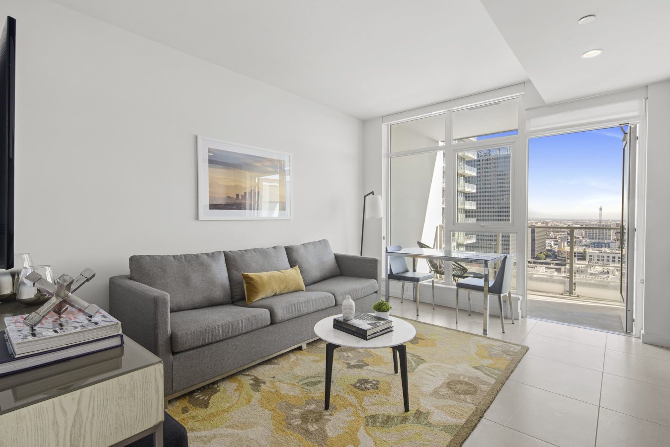 Modern living room with a gray sofa, yellow pillow, round coffee table, and patterned rug. Large windows let in natural light and provide a city view. A small dining table sits near the windows.