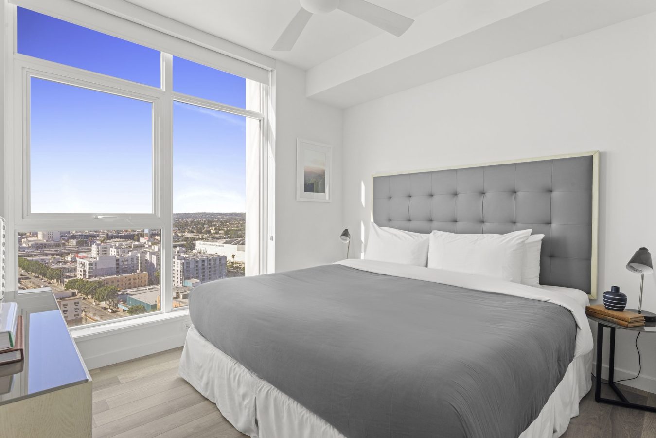 A modern bedroom with a large bed, gray headboard, white bedding, bedside tables with lamps, and a window offering a cityscape view under a clear blue sky.