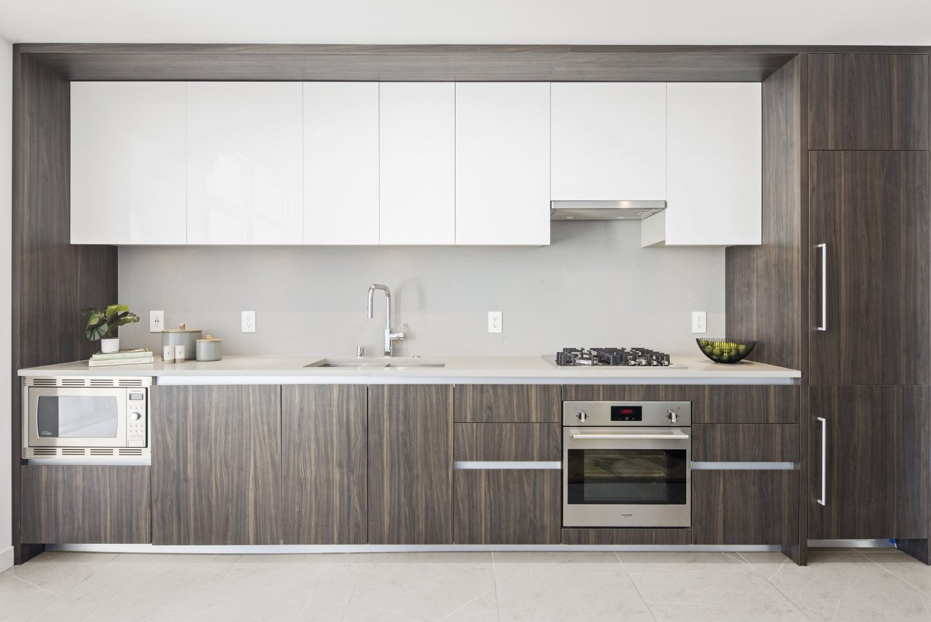 Modern kitchen with wood-grain lower cabinets, white upper cabinets, built-in oven, stovetop, microwave, sink, and minimalist decor, featuring a bowl of green apples and small potted plants.