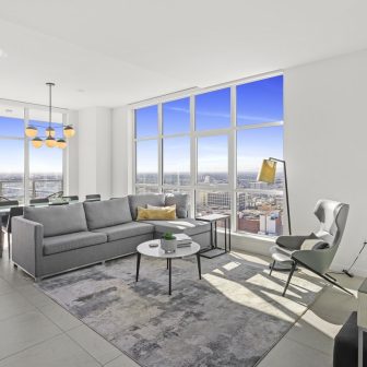 Modern living room with large floor-to-ceiling windows, gray sofa, accent chair, floor lamp, and city skyline view. A rug and coffee table are centered in the sunlit, minimalist space. Dining area visible in the background.