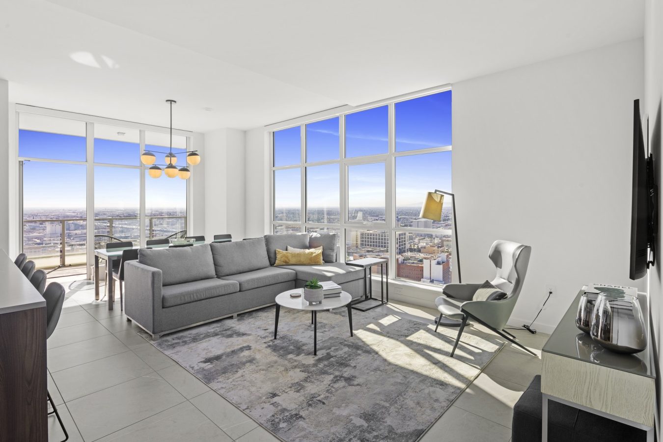 Modern, bright living room with floor-to-ceiling windows showcasing a city view. Furnished with a gray sofa, armchair, rug, TV, coffee tables, and a dining area with a chandelier. Sunlight fills the space.