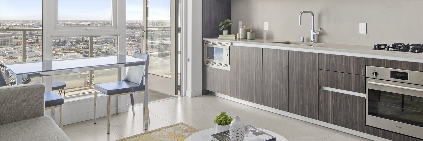 Modern apartment kitchen with wood cabinets, built-in appliances, and a dining table with two blue chairs near large windows offering a city view. Natural light fills the space.