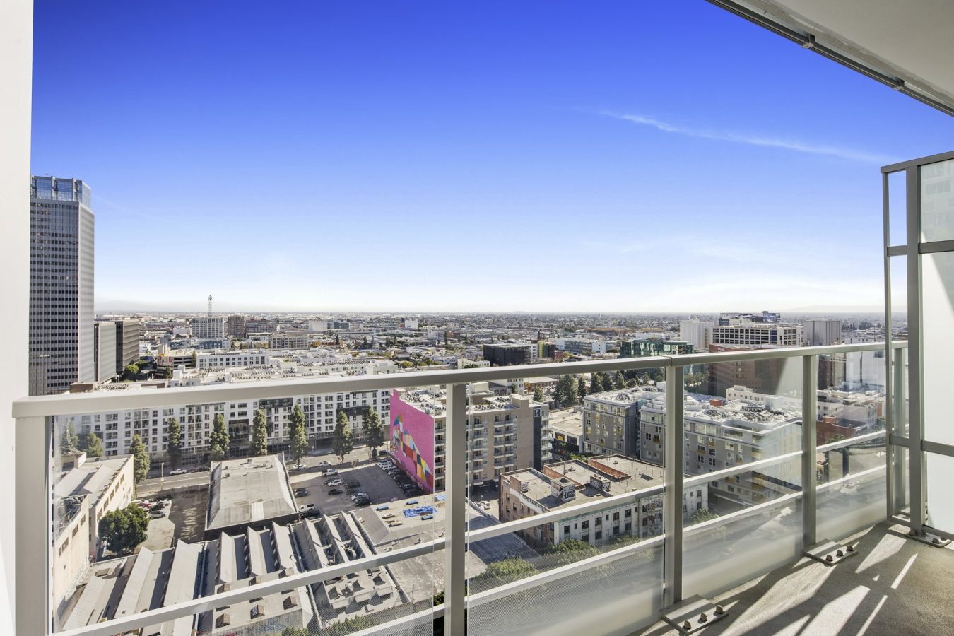 High-rise balcony view overlooking a cityscape with modern buildings, a bright mural on one structure, clear blue sky, and sunlight casting shadows on the glass balcony railing.