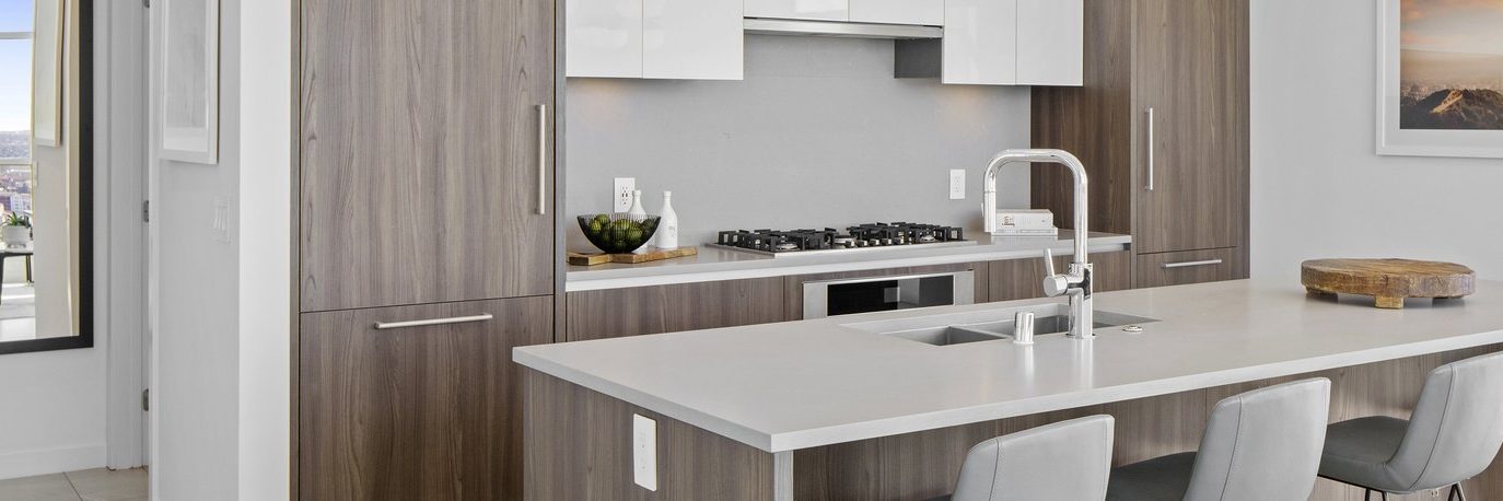 Modern kitchen with wood cabinets, white countertops, built-in oven, gas cooktop, stainless steel faucet, and gray upholstered barstools at a central island. A bowl of fruit and cutting board are on the counters.
