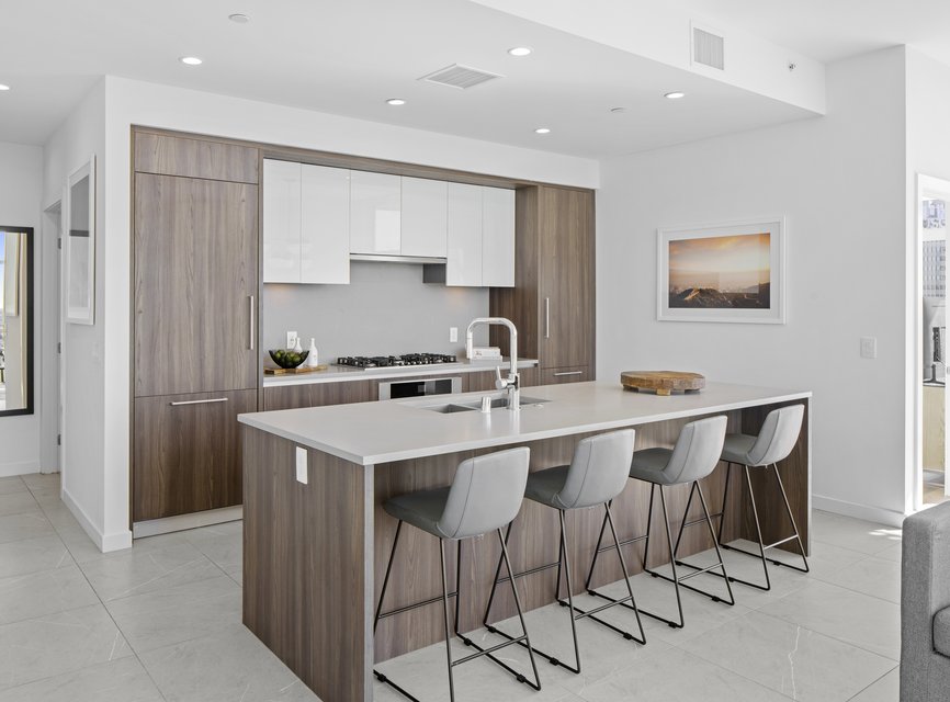 Modern kitchen with wood cabinets, white countertops, a center island with four gray barstools, built-in appliances, and a framed landscape photo on the wall. The space is bright with natural light.