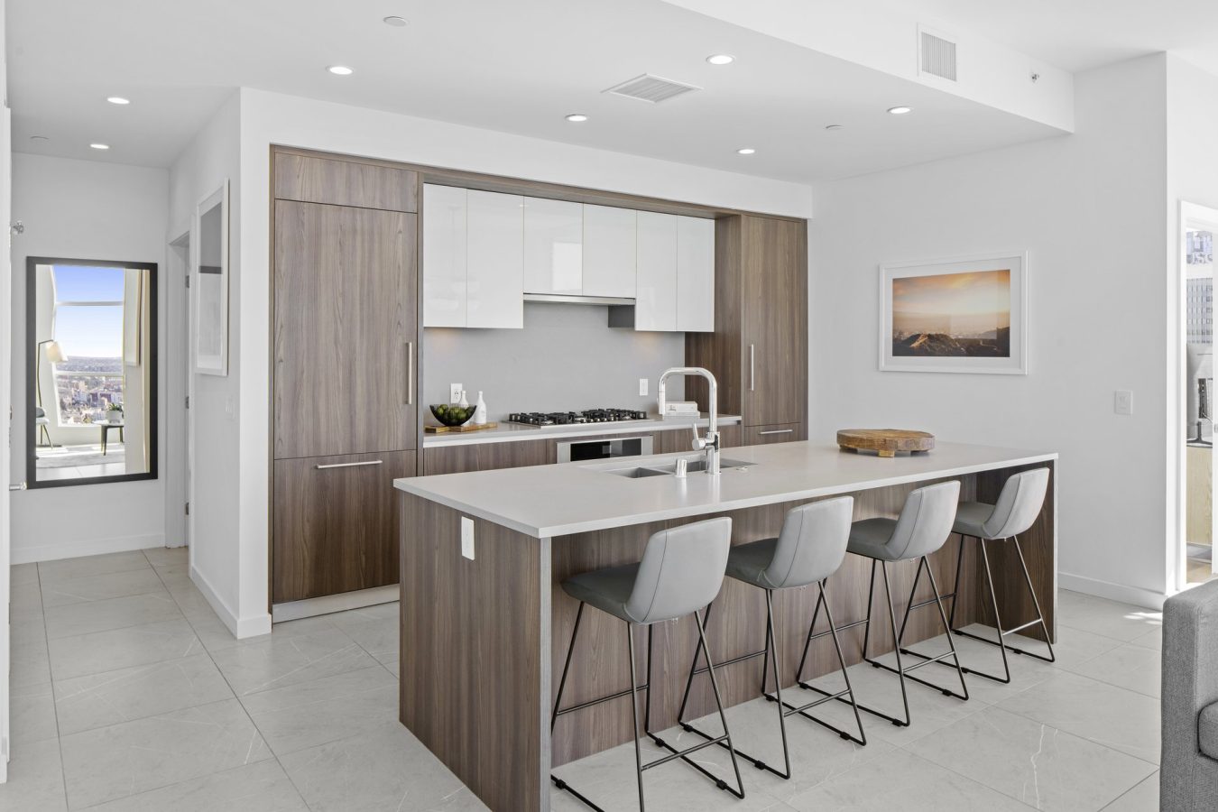Modern kitchen with wood cabinetry, white countertops, a large island with four gray barstools, built-in appliances, and minimalist decor. A window and framed photo hang on the white walls, with city views visible through a doorway.