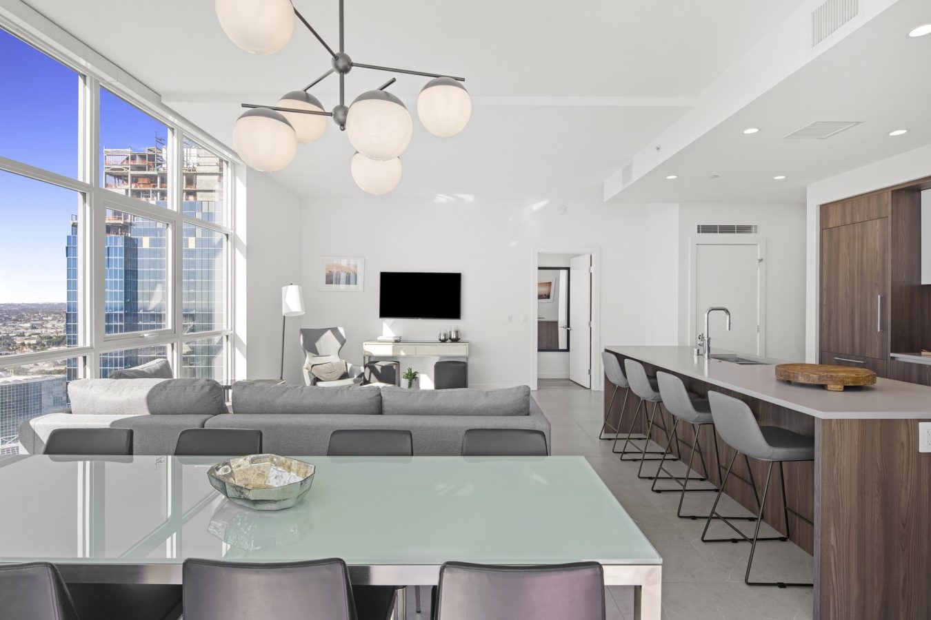 Modern, bright apartment with floor-to-ceiling windows, gray sofa, glass dining table, open kitchen with bar stools, and city view; pendant light fixture hangs from the ceiling.