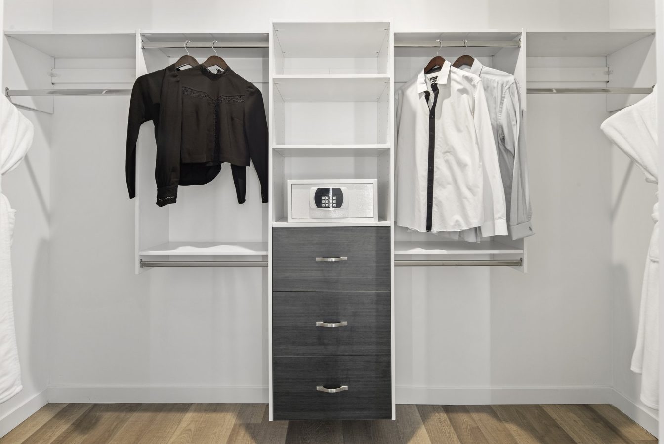 A modern walk-in closet with hanging black and white shirts, open shelves, a digital safe, and dark drawers on a wooden floor. White walls and robes are visible on either side.