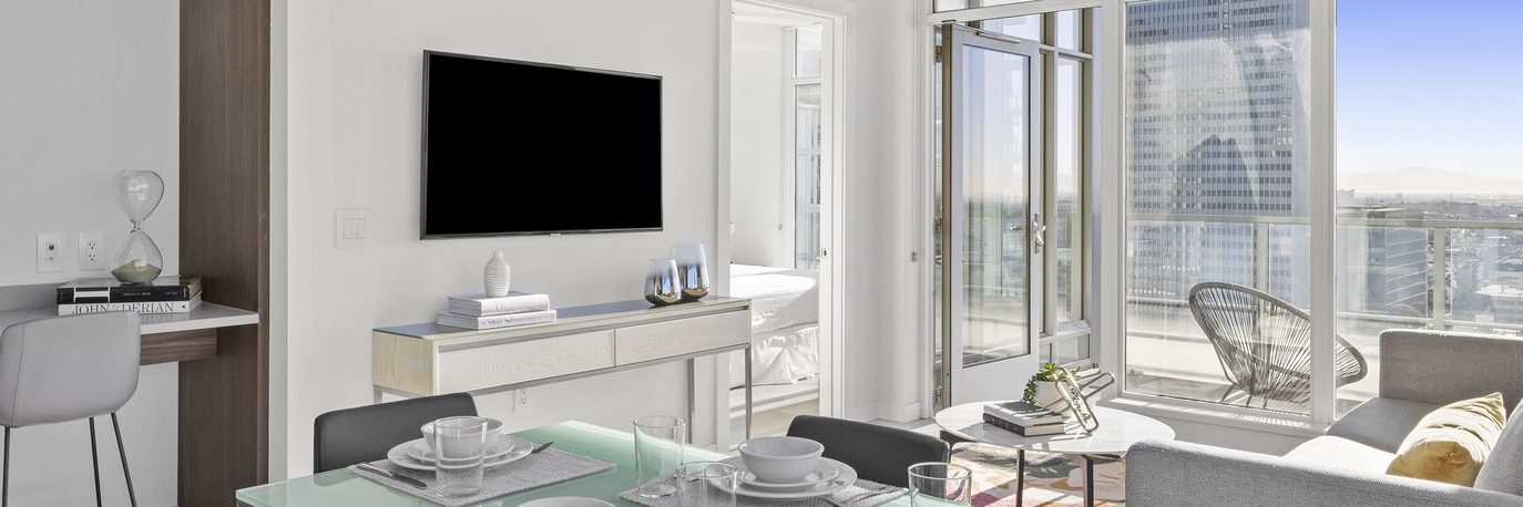 Modern apartment living room with glass dining table, grey sofa, wall-mounted TV, and large windows leading to a balcony with city views. Light decor and natural sunlight fill the space.