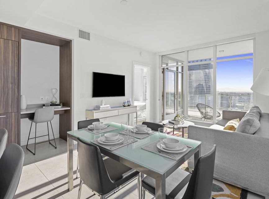 Modern, bright apartment with a glass dining table set for four, a cozy living area, TV, balcony with city views, and a small desk workspace to the left. Light flows in through large windows and a glass door.
