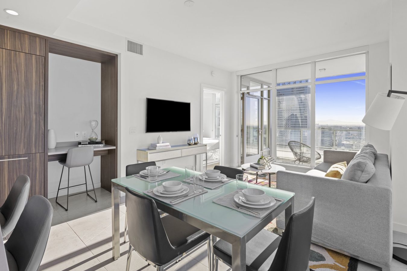 Modern, bright apartment interior with a dining table set for four, gray sofa, wall-mounted TV, small workspace, and large windows leading to a balcony with city views and blue sky outside.