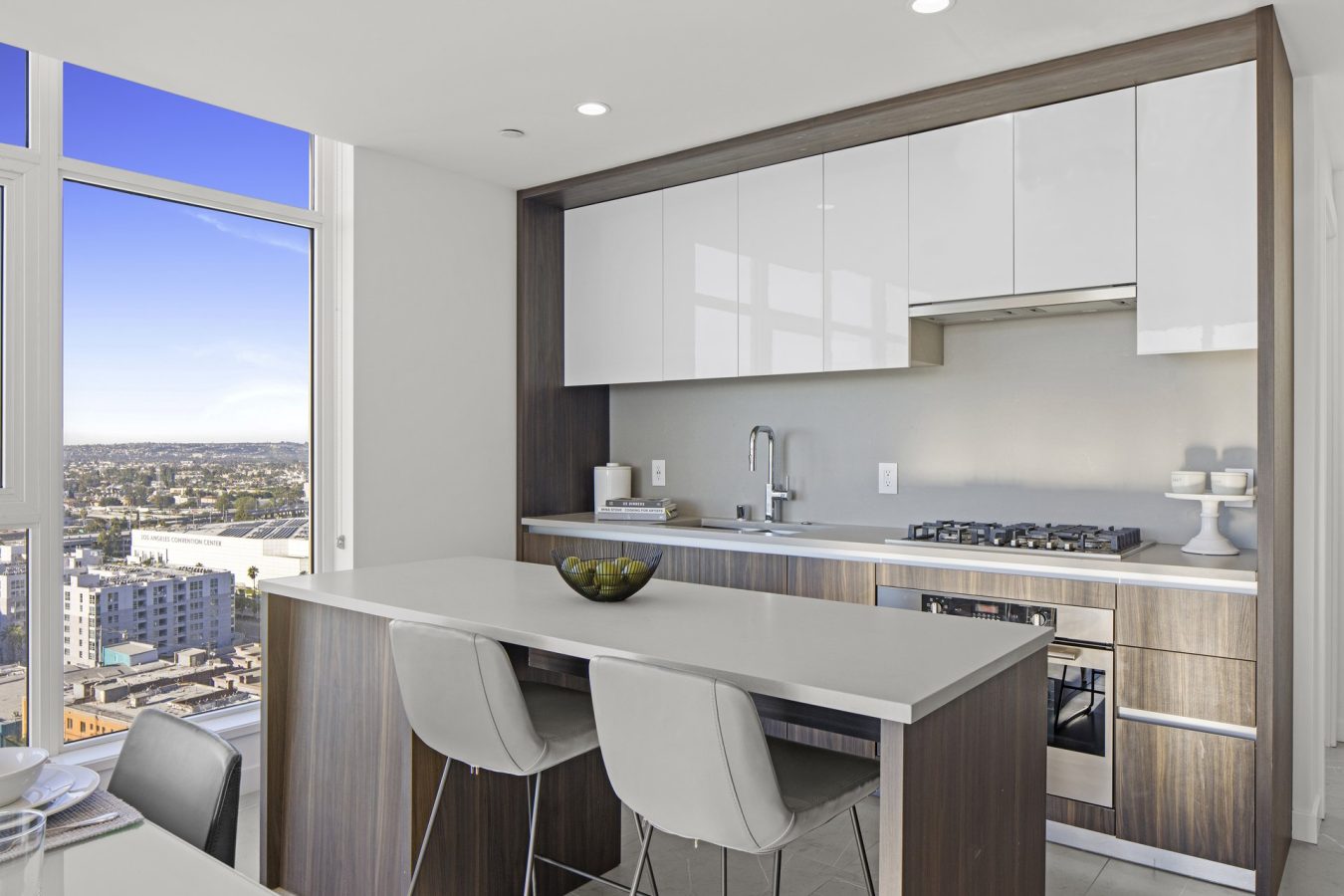 Modern kitchen with wood and white cabinetry, a built-in oven, gas stove, and an island with two gray chairs. Large window offers a cityscape view under a clear blue sky. Bowl of green apples on the counter.