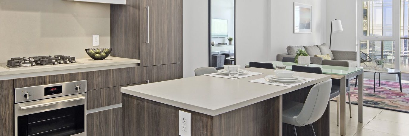 Modern kitchen with wood cabinets, stainless steel oven, and gas stove, connected to a dining area and living room with gray sofa, glass table, and large windows letting in natural light.