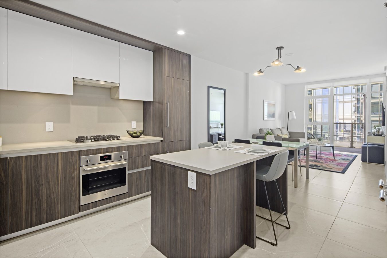 Modern open-plan kitchen and living area with wood cabinets, built-in oven, small island with two bar stools, dining table, sofa, and large windows letting in natural light.