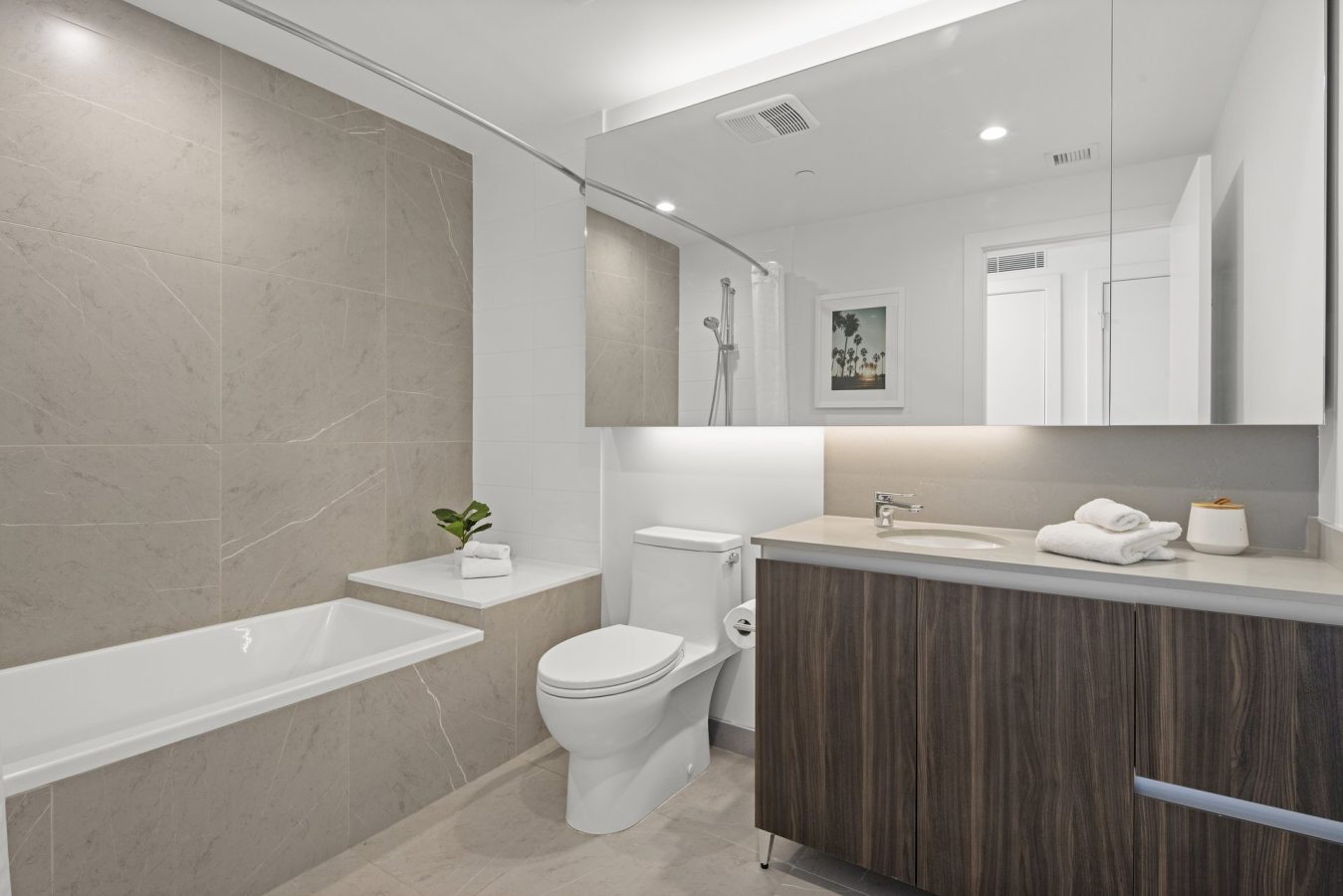 Modern bathroom with a bathtub, toilet, and sink vanity with wood cabinetry. Large wall tiles, a potted plant, white folded towels, and a large mirror create a clean and minimalist look.