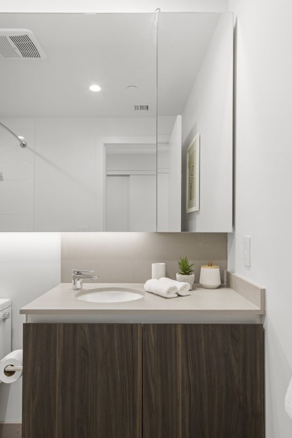 Modern bathroom with a wooden vanity, white countertop, sink, faucet, rolled towels, soap dispenser, and small plant. A large mirror and minimal decor create a clean, bright look.