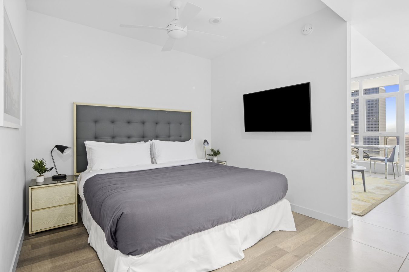 Modern bedroom with a large bed, gray tufted headboard, two nightstands with lamps and plants, wall-mounted TV, ceiling fan, and view into a bright living area with cityscape visible through window.