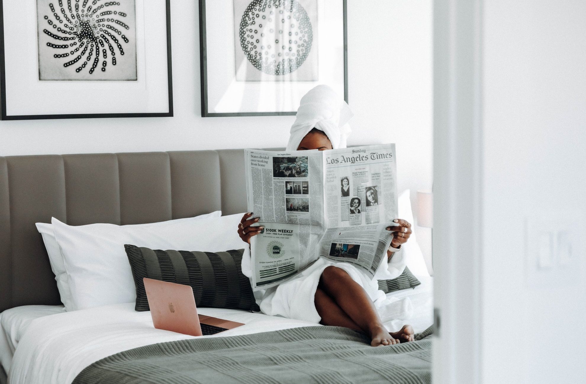A person in a white bathrobe and towel sits on a bed, reading the Los Angeles Times newspaper. A rose gold laptop is beside them on the bed, and modern art hangs on the wall behind them.