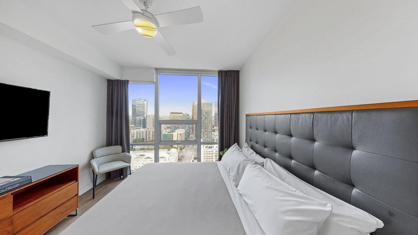 Modern bedroom with a large bed, padded headboard, wall-mounted TV, armchair, ceiling fan, and a large window offering a city skyline view. Room features neutral tones and minimal decor.