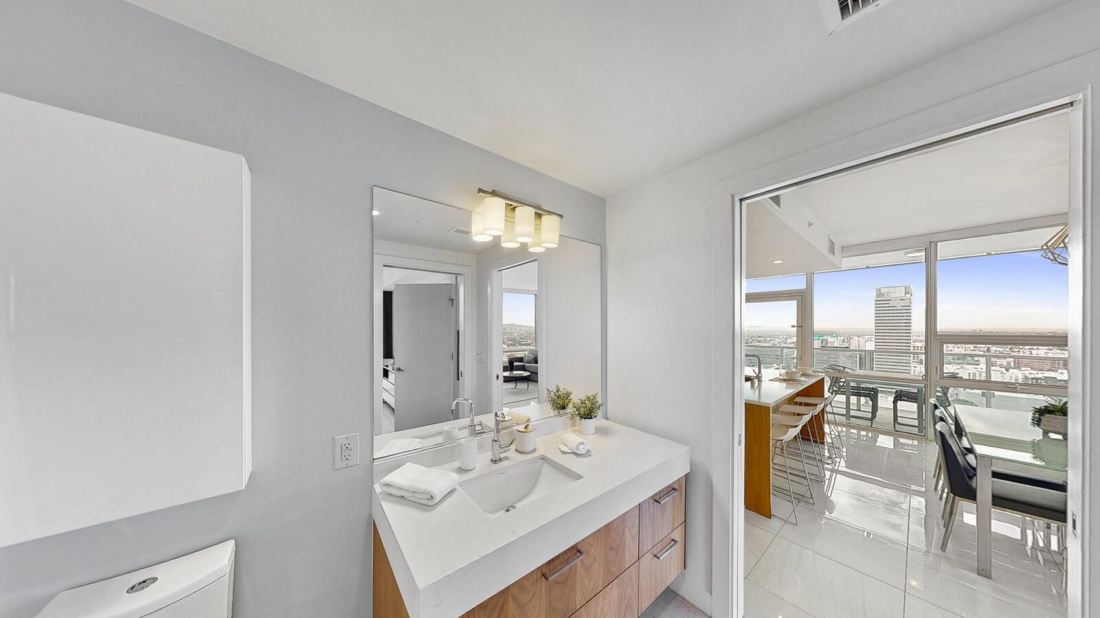 Modern bathroom with double sinks and large mirror, opening to a bright kitchen and dining area with floor-to-ceiling windows and city views in the background.