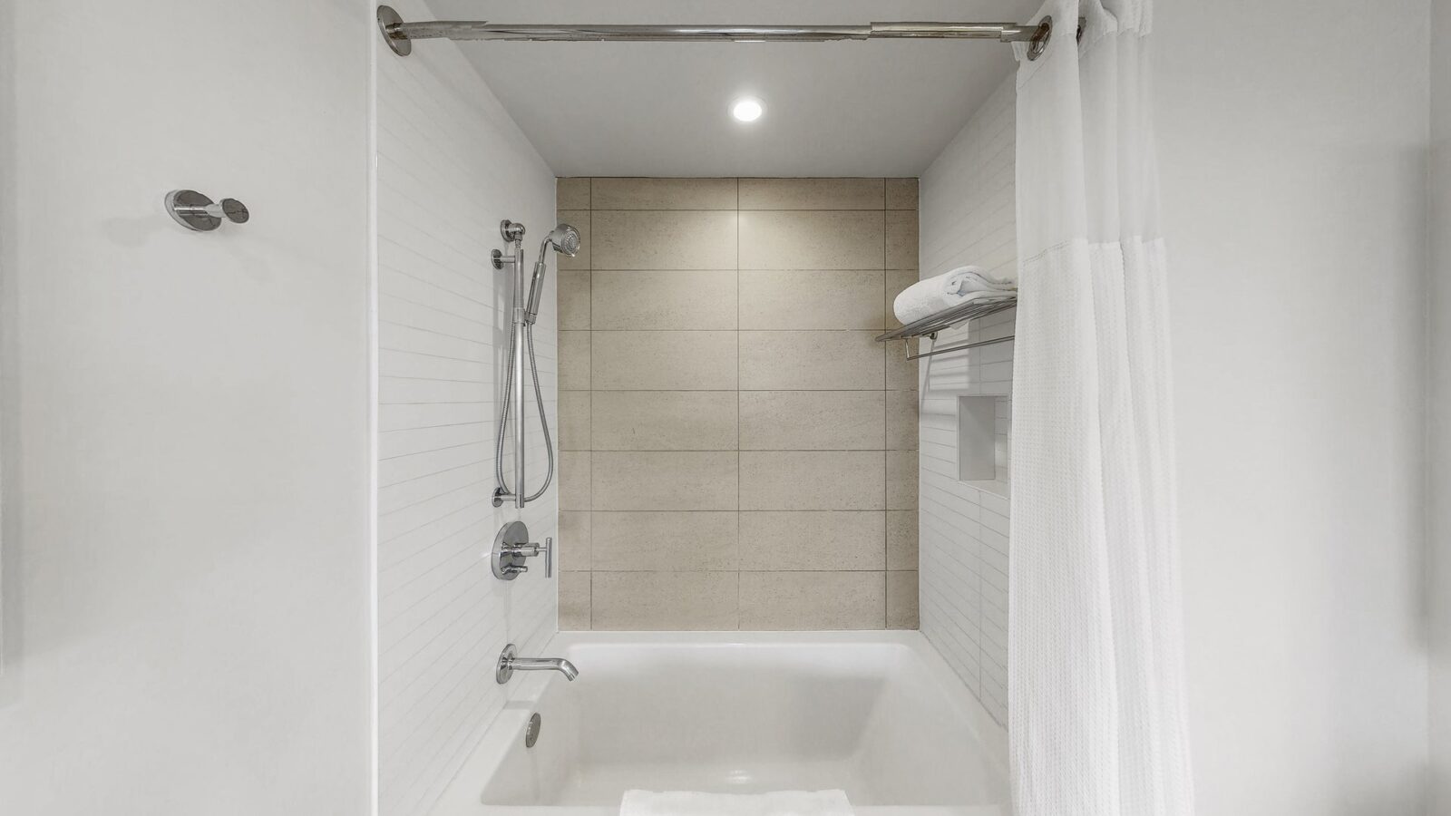 A modern, clean bathroom with a white bathtub, tiled walls, silver shower fixtures, a white shower curtain, a towel bar with folded towels, and a built-in shelf under bright overhead lighting.