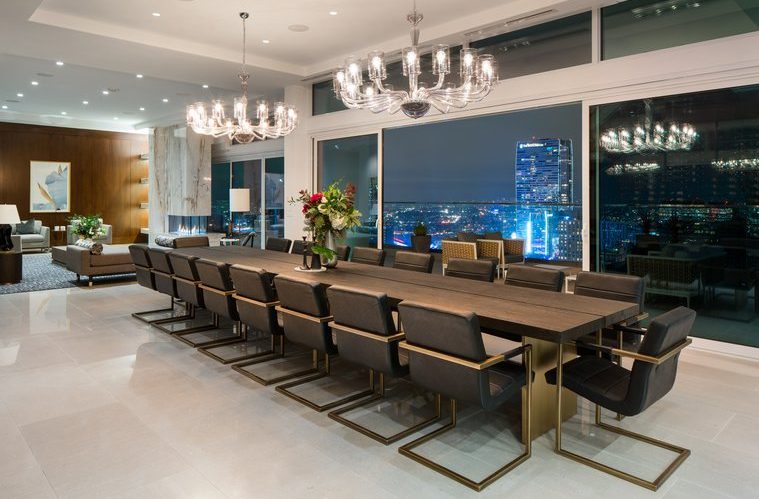 A modern dining room with a long wooden table, twelve black cushioned chairs, two large chandeliers, flowers as the centerpiece, and floor-to-ceiling windows overlooking a city skyline at night.