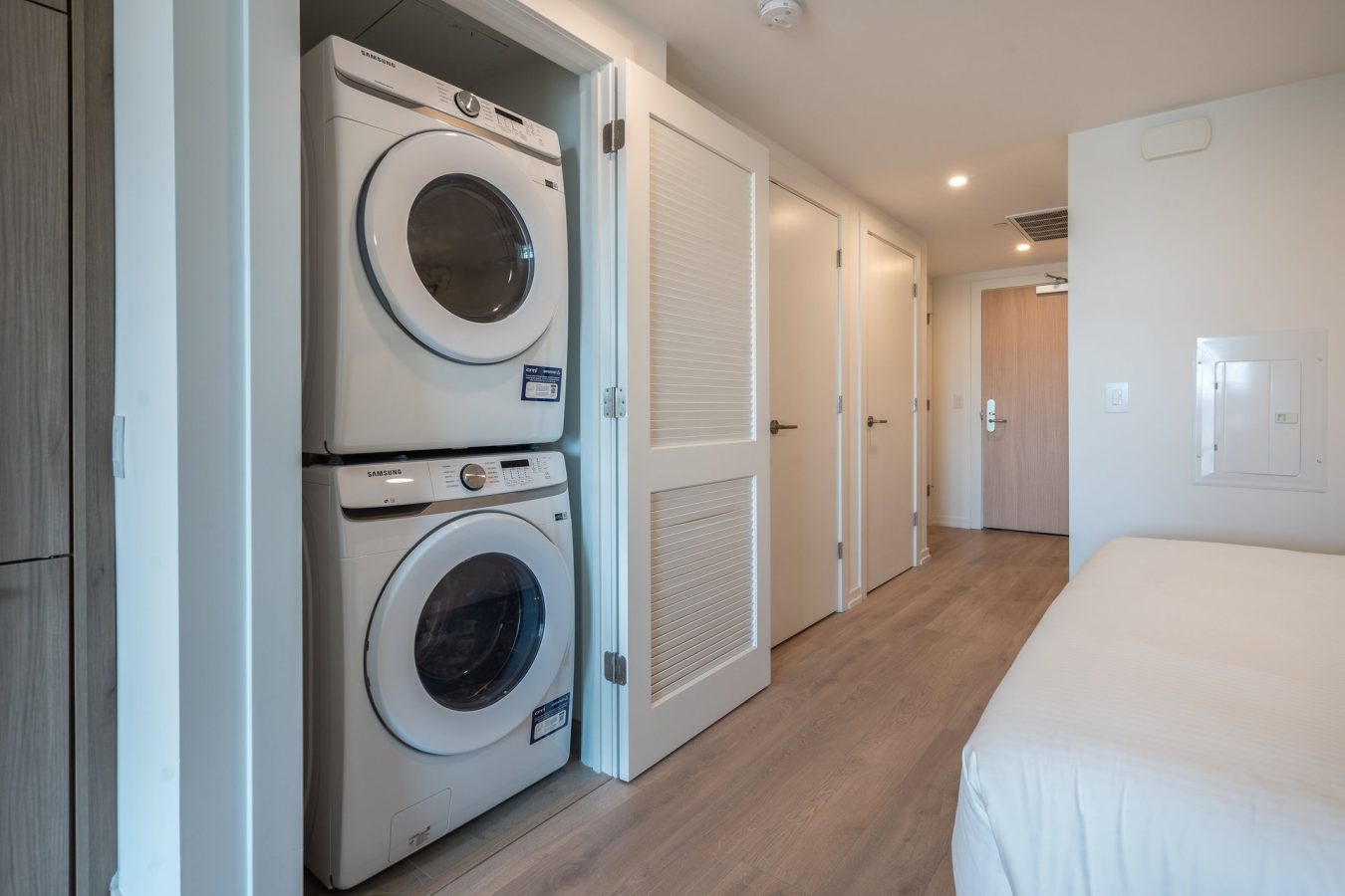 A stacked washer and dryer set is installed in a closet next to a white door in a modern, bright room with light wood flooring. A neatly made bed is partially visible in the foreground.