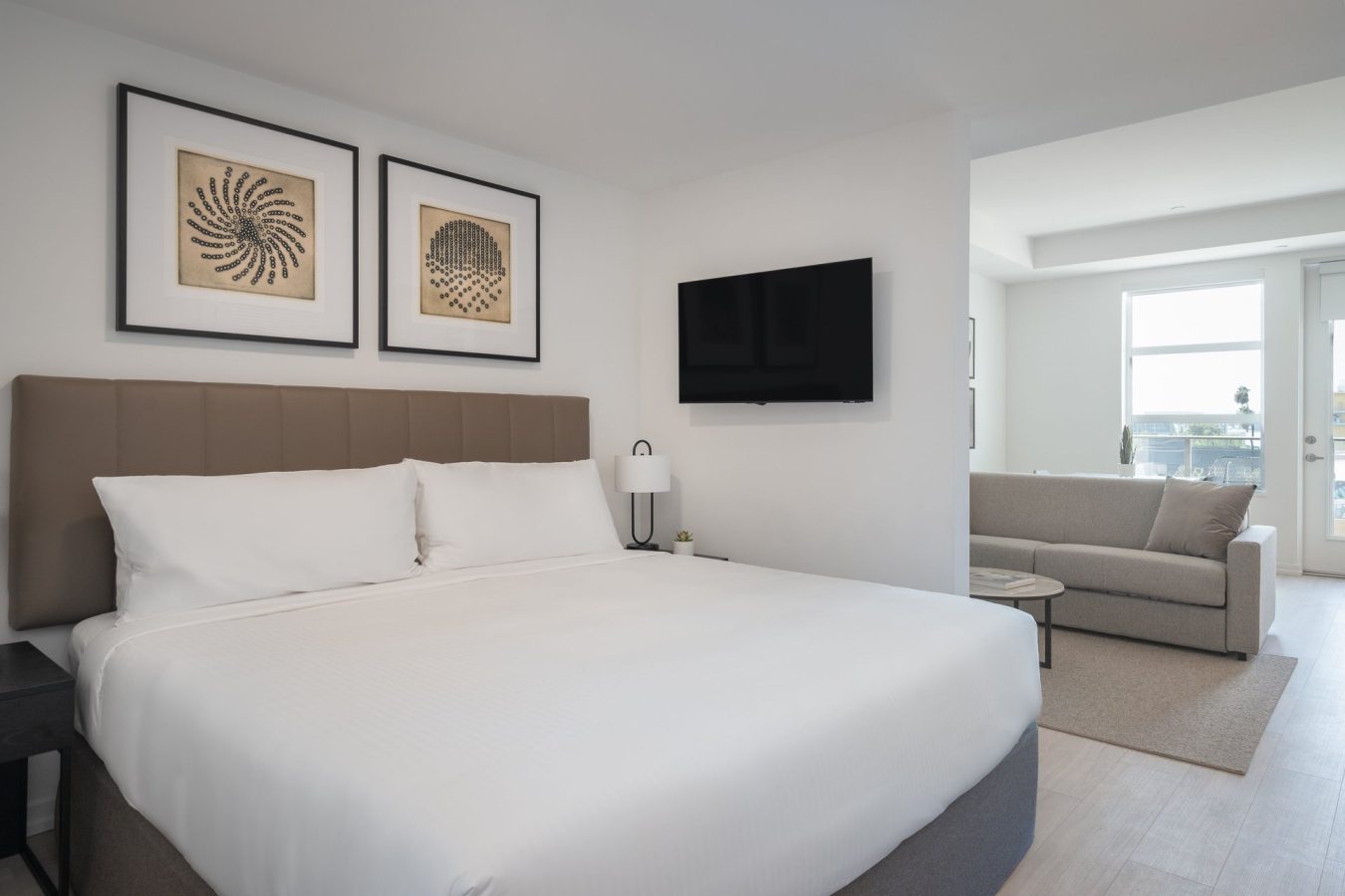 A modern hotel room with a large bed, white bedding, two framed artworks above the headboard, a wall-mounted TV, a small lamp, and a sitting area with a light gray sofa by a window.