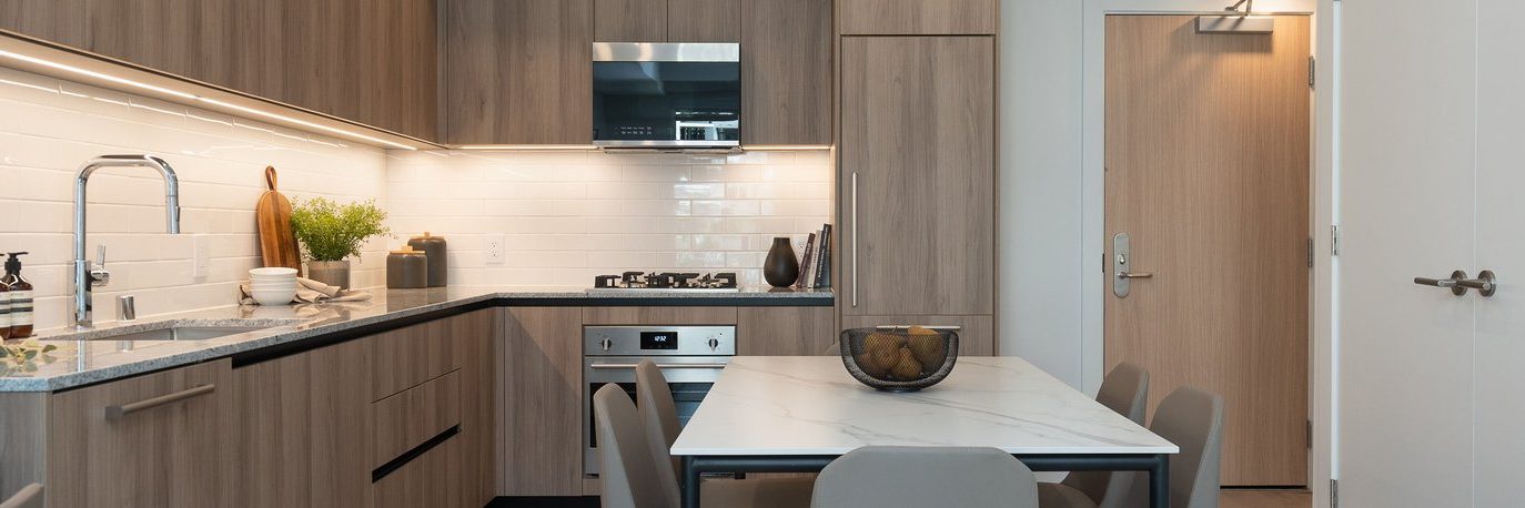 Modern kitchen with light wood cabinets, built-in appliances, white tile backsplash, and a marble dining table with four gray chairs. A door is visible in the background, and the space is tidy and well-lit.
