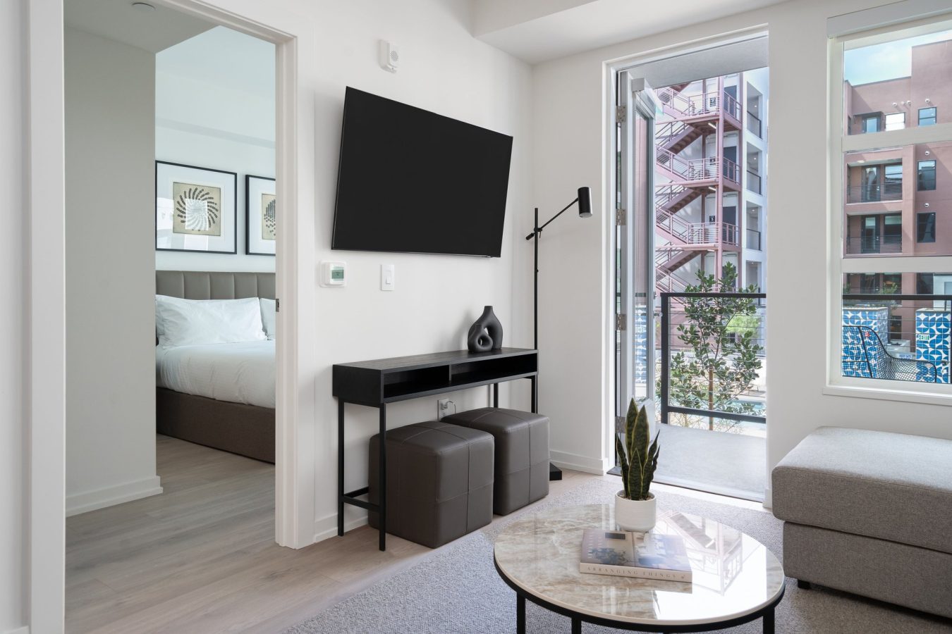 Modern apartment living room with a wall-mounted TV, console table, two ottomans, and a round coffee table with a plant. Bedroom visible through an open door; large windows show an outdoor fire escape and buildings.