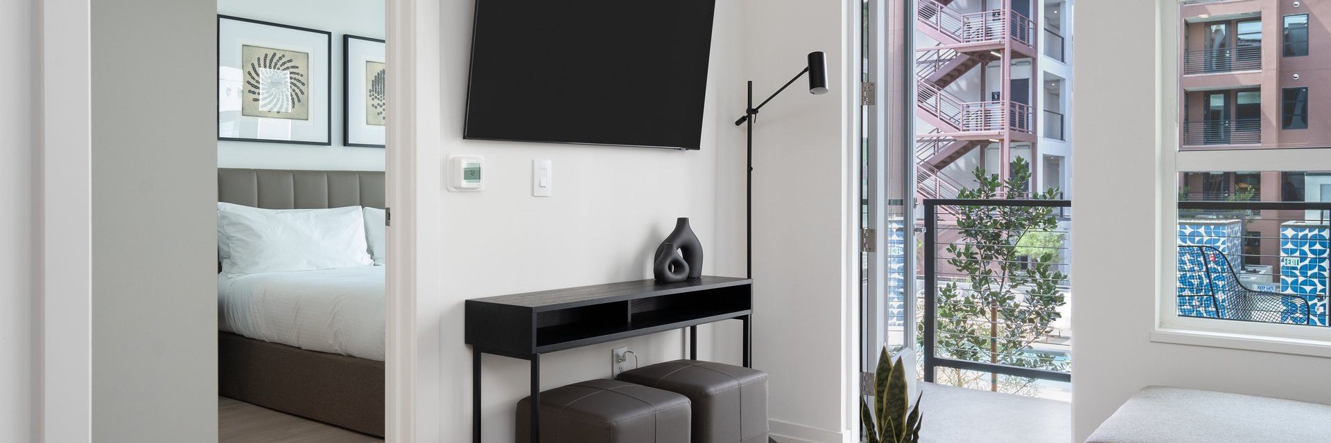 Modern apartment interior with a wall-mounted TV above a black console table, two ottomans, a floor lamp, and a balcony view. A bedroom with a neatly made bed is visible through an open door.