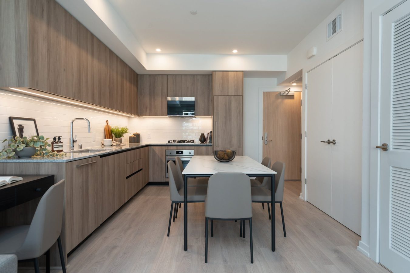 Modern kitchen with light wood cabinets, white countertops, built-in appliances, and a dining table with four gray chairs. The space is well-lit with both ceiling and under-cabinet lights.