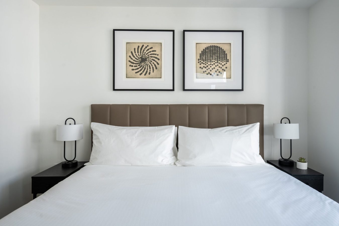 A neatly made bed with white linens, two white pillows, and a beige padded headboard. Two framed abstract artworks hang above the bed, flanked by black bedside tables with matching lamps and a small plant.