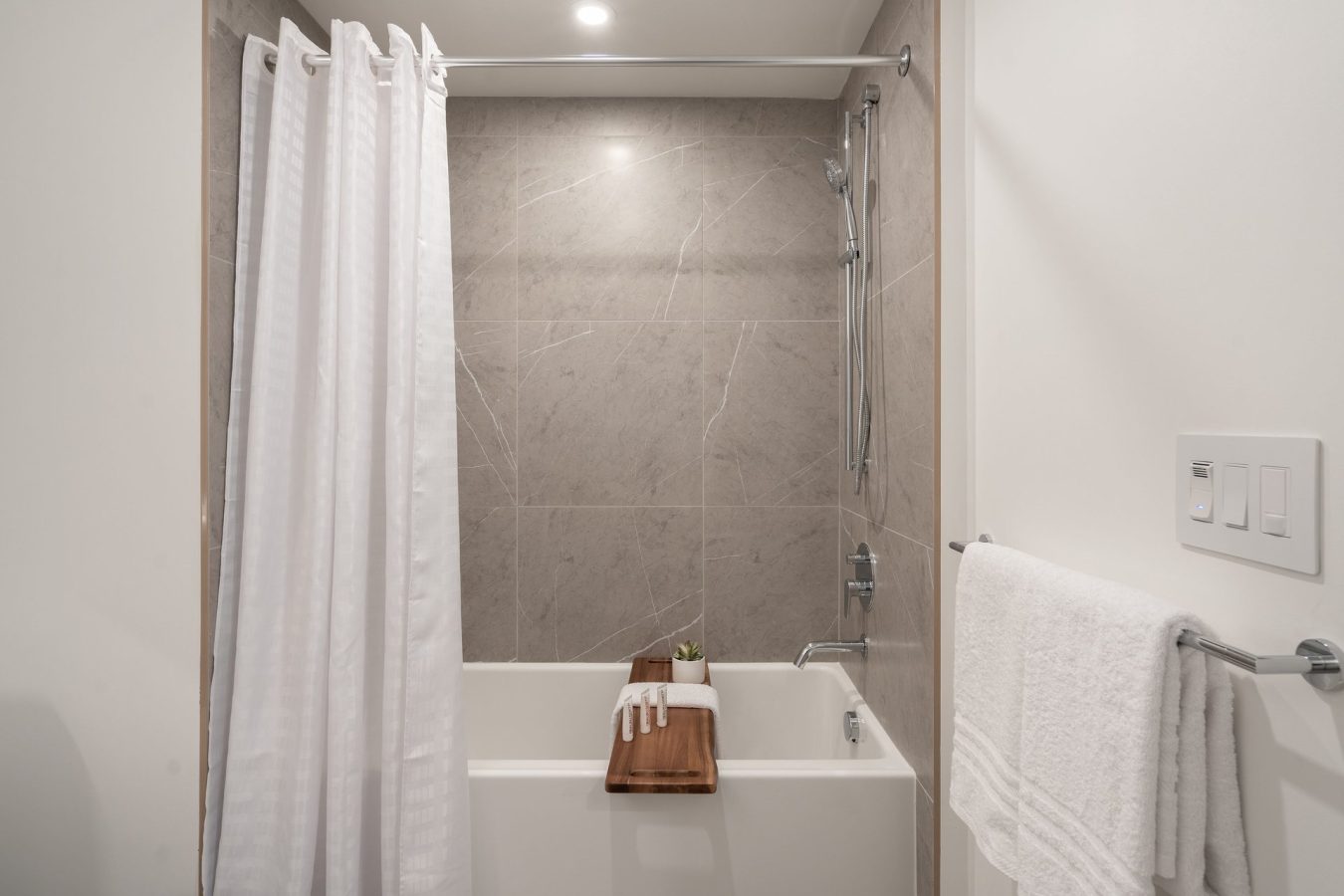 A modern bathroom featuring a white bathtub with a gray tiled wall, a white shower curtain, a towel bar with a white towel, and a wooden bath tray holding a small plant and folded white towels.