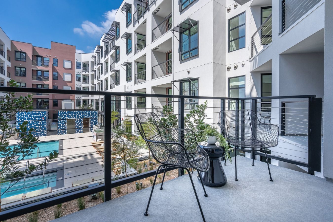 Modern apartment balcony with two black chairs and a small table, overlooking a courtyard with a swimming pool, plants, and other apartment buildings under a blue sky.