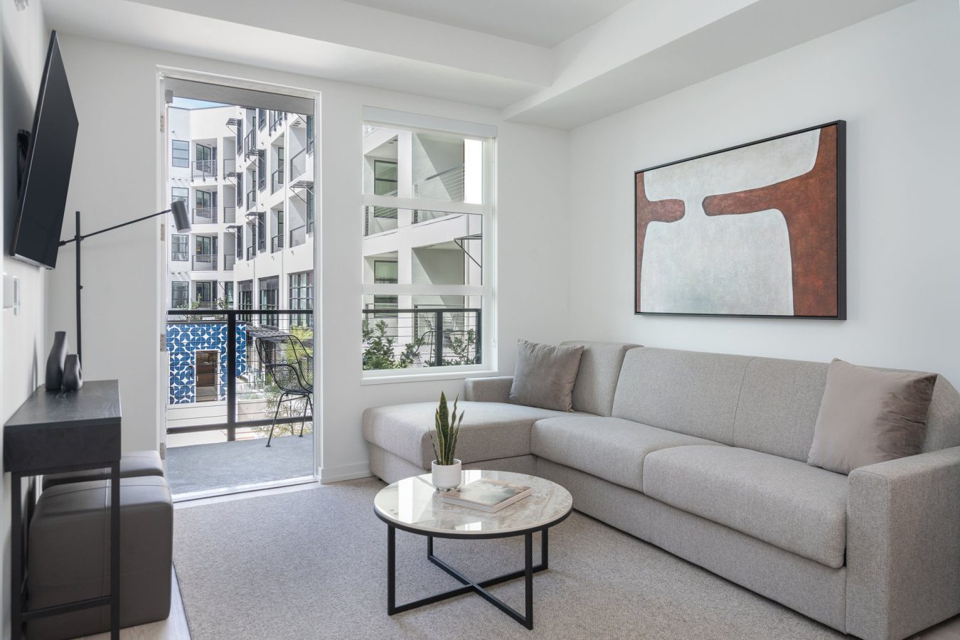 A modern living room with a light gray sectional sofa, abstract wall art, a round marble coffee table with a plant, a wall-mounted TV, and large windows letting in natural light with a view of a balcony and nearby buildings.