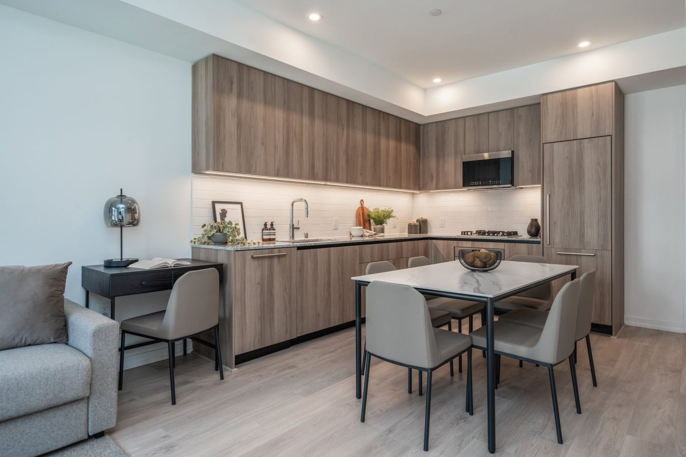 Modern kitchen with light wood cabinets, built-in appliances, marble dining table with four beige chairs, and a small black desk beside a light gray sofa. The space is bright with recessed ceiling lights.