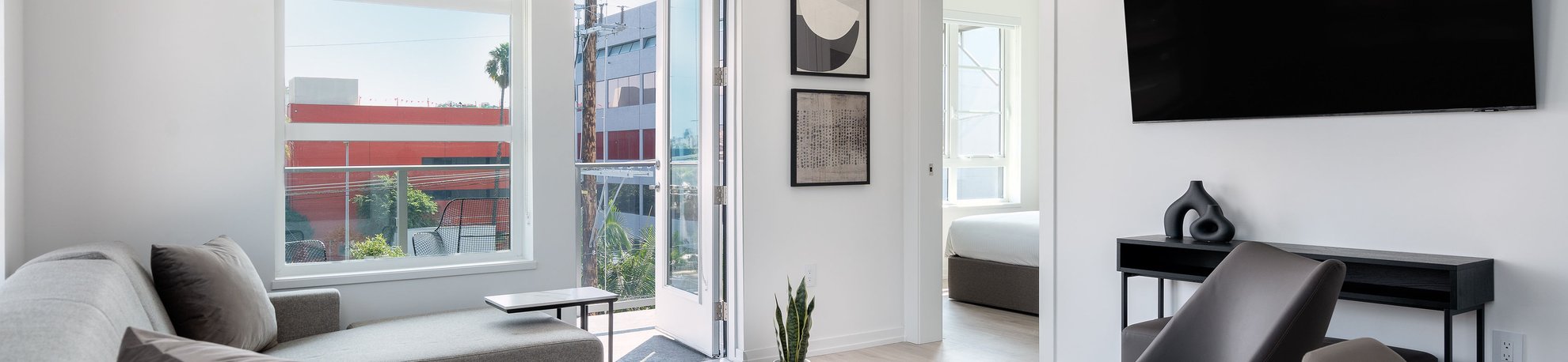 Modern living room with a gray sofa, coffee table, wall-mounted TV, and dining area. Large windows and a glass door provide natural light, with a bedroom visible through an open doorway. Minimalist decor throughout.