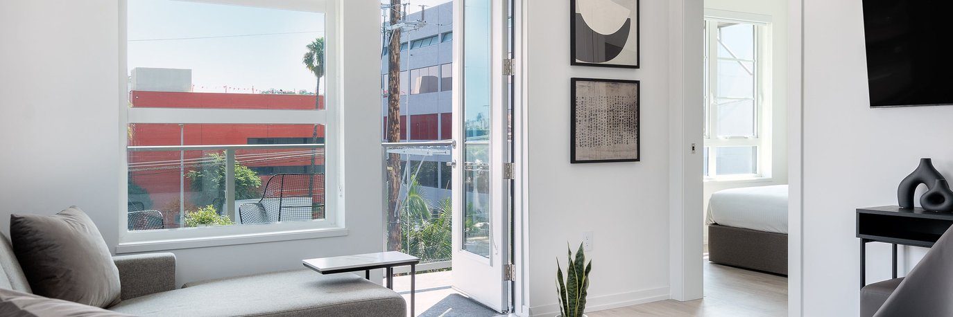 A bright, modern living room with large windows, a gray sectional sofa, a small table, and a potted plant. An open door leads to a bedroom with a white bed and more windows; minimalist art hangs on the wall.