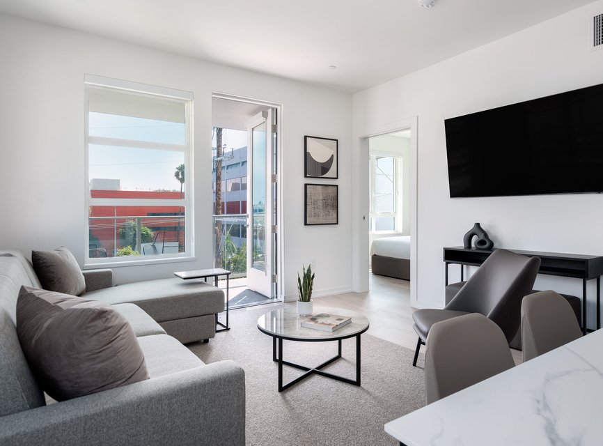 Modern living room with gray sofas, a round coffee table, wall-mounted TV, minimalist decor, and large windows letting in natural light. A bedroom is visible through an open door and a balcony can be seen outside.