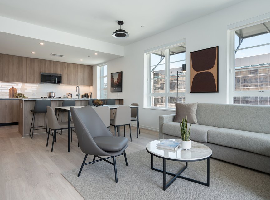 A modern open-plan living room and kitchen with light wood floors, a gray sofa, armchair, round coffee table with a plant, dining table, and minimalist decor. Large windows let in natural light.
