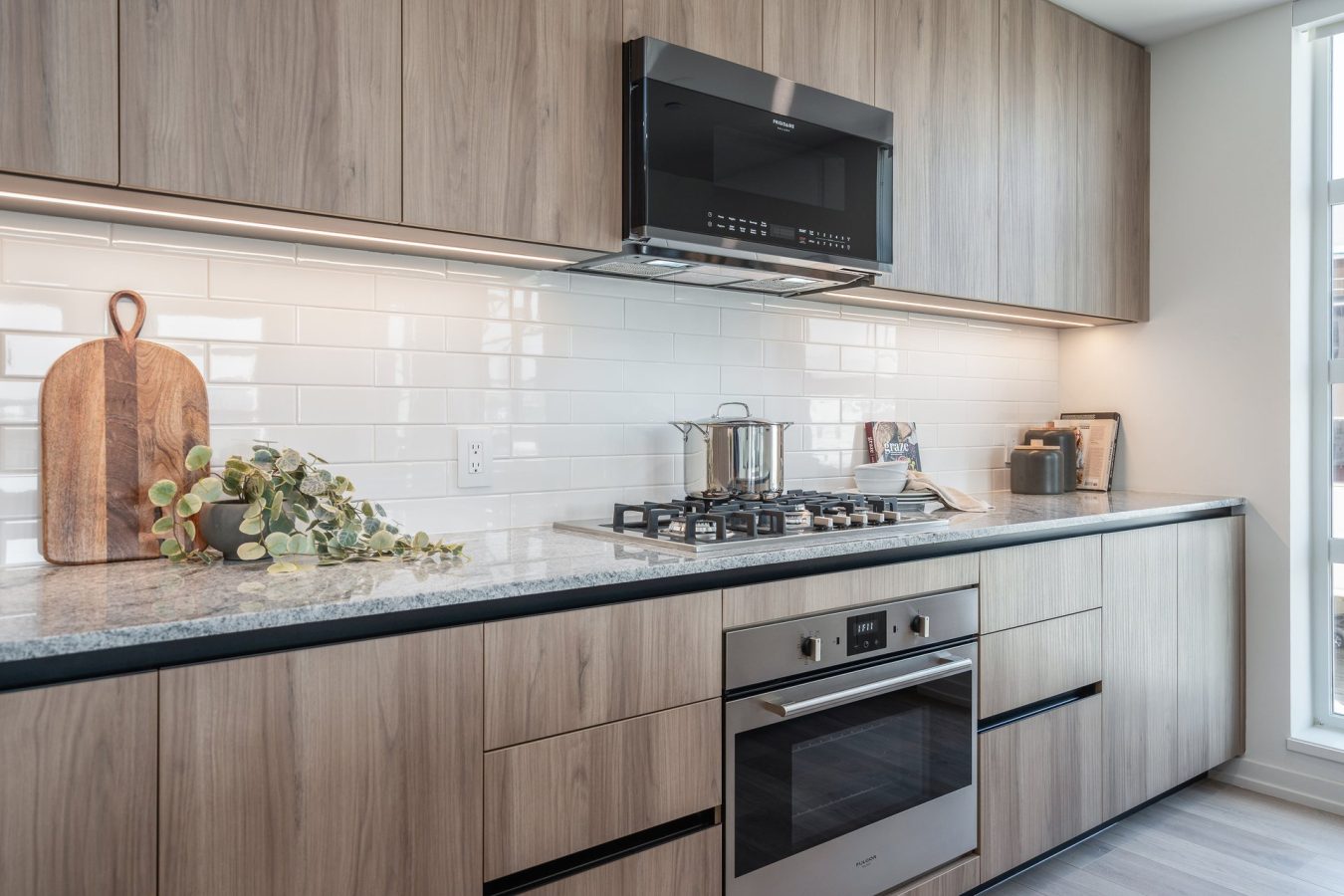 Modern kitchen with light wood cabinets, white subway tile backsplash, stainless steel oven, gas cooktop, pot on the stove, potted plant, cutting board, and sunlight streaming through a large window.