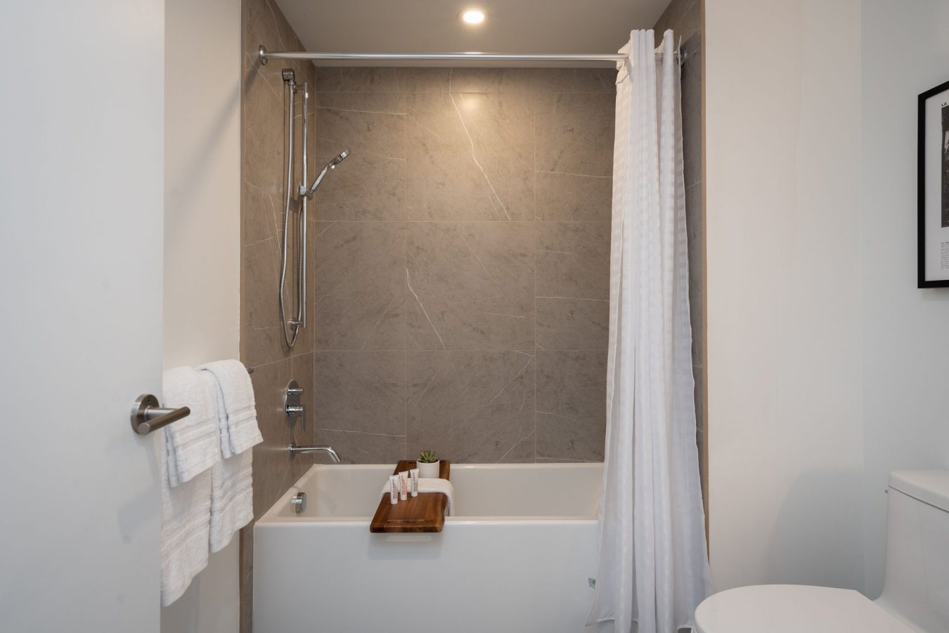 Modern bathroom with a white bathtub, gray tiled wall, showerhead, white shower curtain, white towels on a rack, and a wooden bath tray holding towels and a small plant. Part of a toilet is visible on the right.
