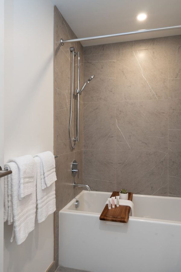 Modern bathroom with gray tile walls, a white bathtub, a mounted showerhead, white towels on a rack, and a wooden bath tray holding folded towels, toiletries, and a small potted plant.