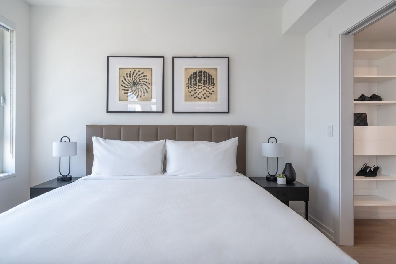 A modern, minimal bedroom with a neatly made bed, white bedding, two bedside tables with lamps, and two framed abstract artworks above the headboard. An open closet with shelves is visible to the right.