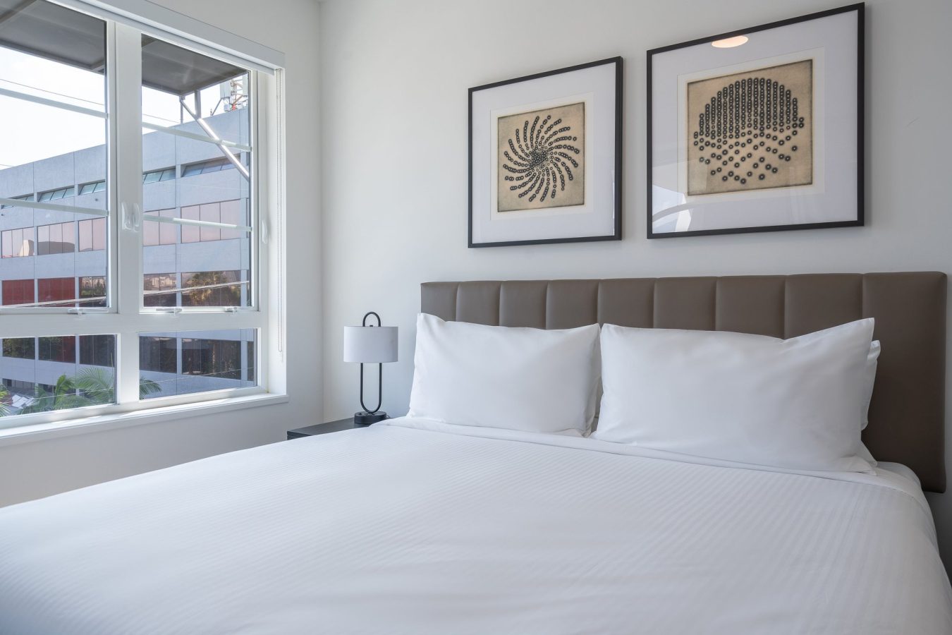 A neatly made bed with white linens sits against a wall with two framed abstract art pieces above the headboard. A window to the left lets in natural light and shows part of a modern office building outside.